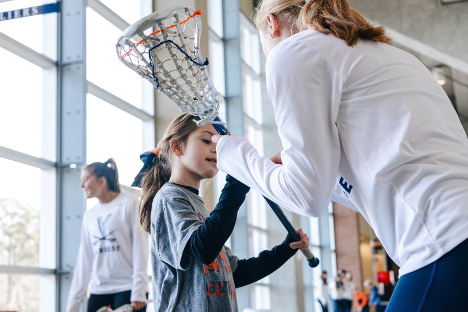A UVA lacrosse team member hands a lacrosse stick to a little girl