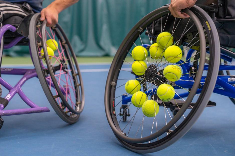 Two wheelchairs, with tennis balls in their spokes, rest on a blue tennis court. 