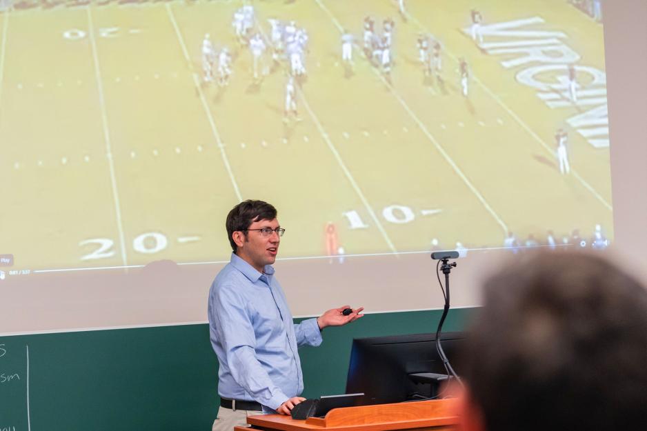 Richard Re lectures from a podium in front of a projection of a football game