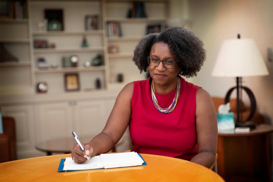 Stephanie Rowley sits at a table writing in a notebook and smiling at the camera