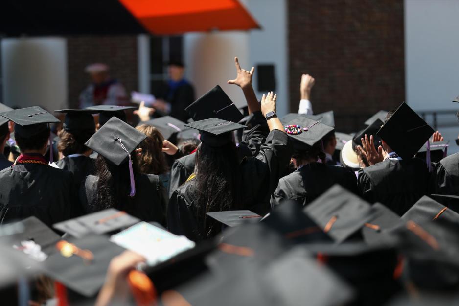 In a crowd of graduating students in caps, one student raises her hand, making the symbol for I love you.