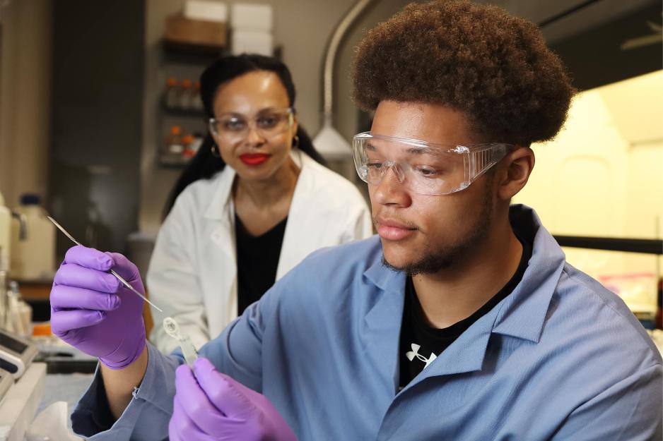 Caleb Tyson sits in the foreground wearing safety goggles, gloves, and a lab coat.