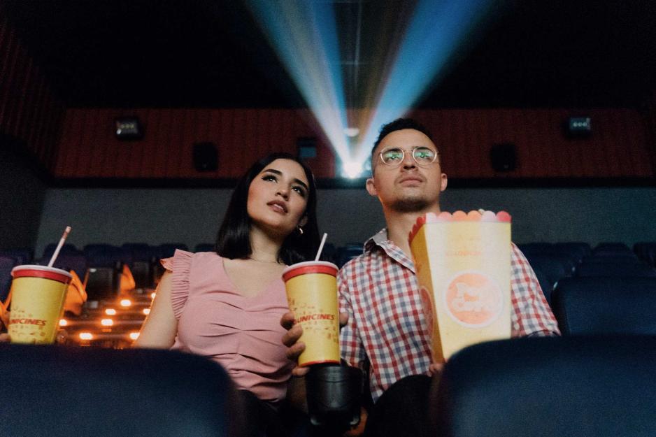 A man and a woman sit in a movie theater, holding drinks and popcorn