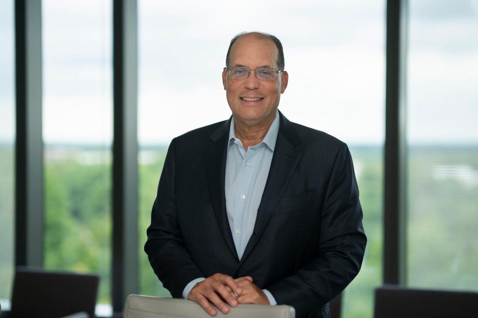 Tom Baltimore stands with his hands on the back of a chair and smiles at the camera in front of the glass window of a conference room