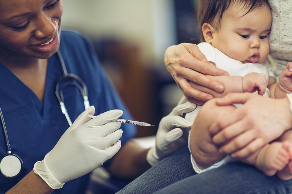 A nurse injects a vaccine into the thigh of a baby sitting in her mother's lap