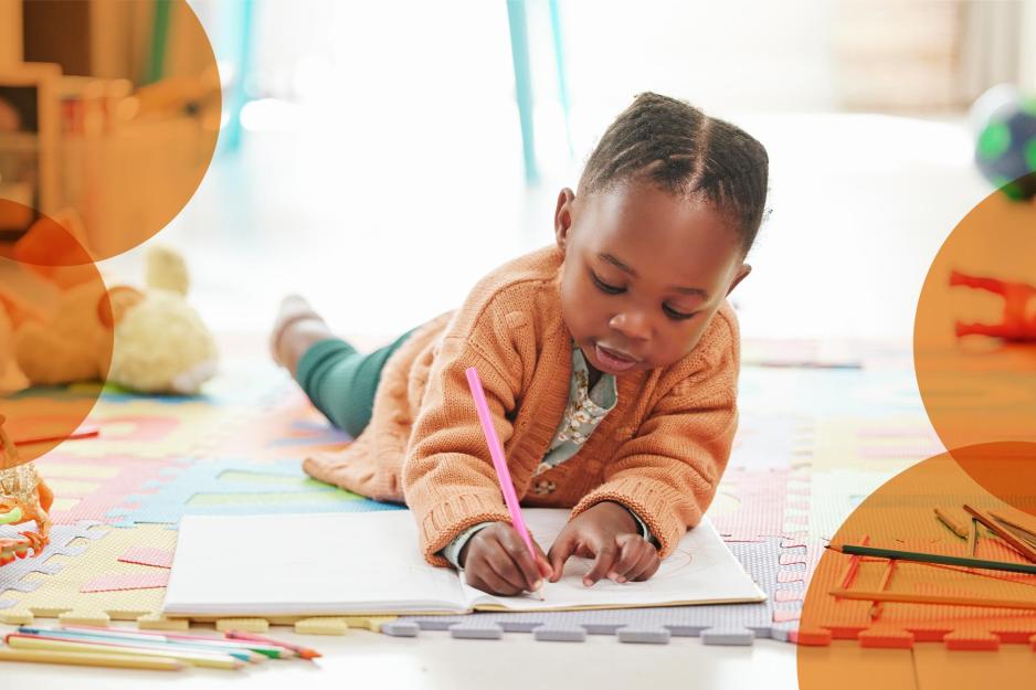 A small child lays on the ground of a preschool classroom, drawing with a pink colored pencil
