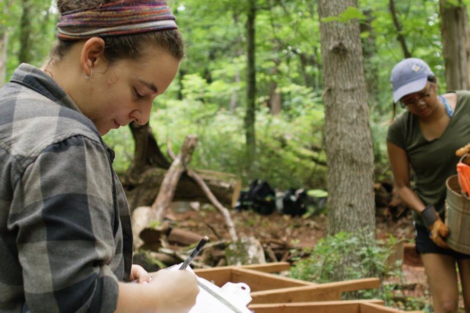 Woman writing on a clipboard while others carry buckets of dirt