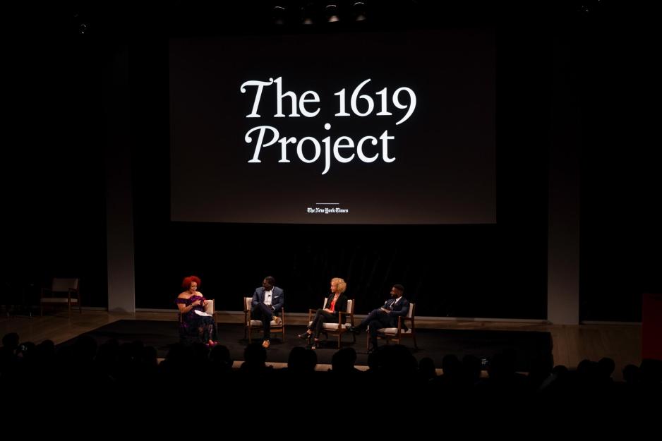 four speakers sit in chairs on a stage speaking with a sign above them that reads The 1619 Project