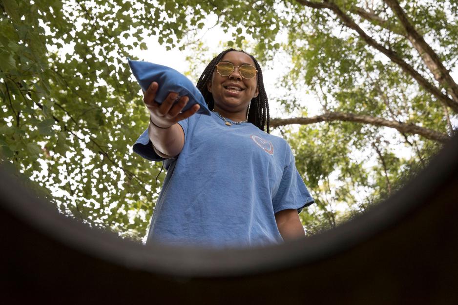 Camera is looking out of the hole to a Student standing over a cornhole boards hole with a bean bag.  