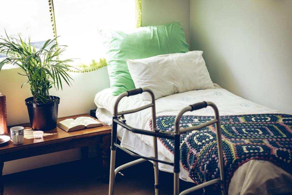 Empty bed at a long-term care facility with a walker next to the bed and a crocheted afghan laying on the bed