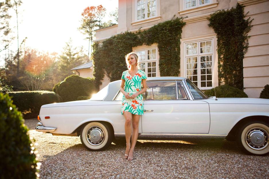 Woman leaning next to a car in a dress.  The dress has palm leaves and orange flowers, has a v-neck and stops about six inches above the knee