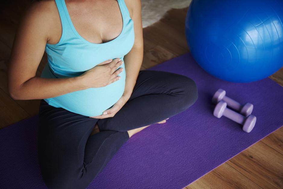 Pregnant woman holding her stomach as she sits on an exercise mat next to hand weights and yoga ball