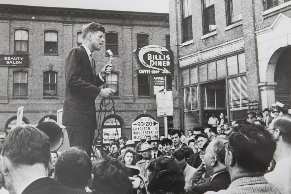 Black and White image of John F. Kennedy standing above the crowd in a street talking into a microphone