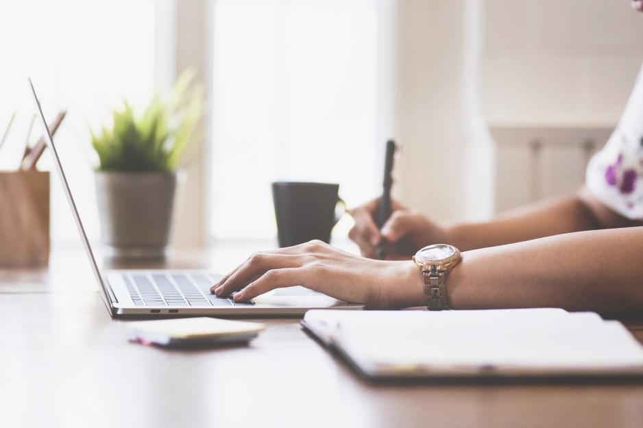 Person sitting at  table with a computer writing in a notebook