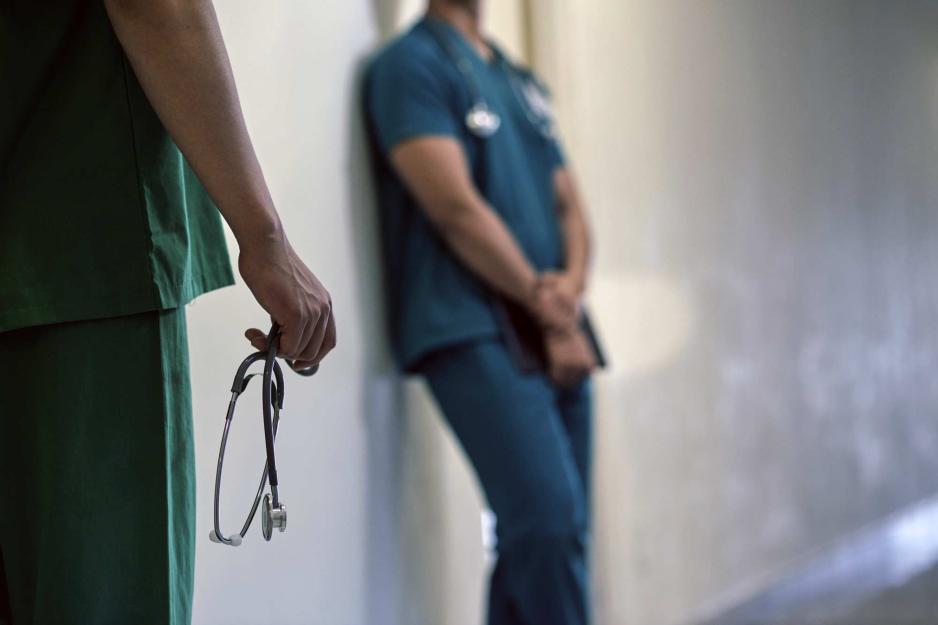 Two medical professionals one leans against a wall and the other stands holding a stethoscope