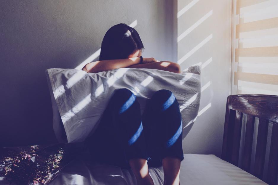 Person sitting on a bed with their knees up and with their head resting on their arms on a pillow and their knees