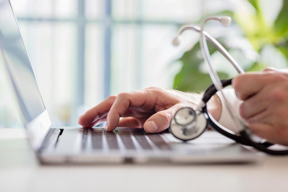 person typing on a laptop with right hand and holding a stethoscope in their left hand