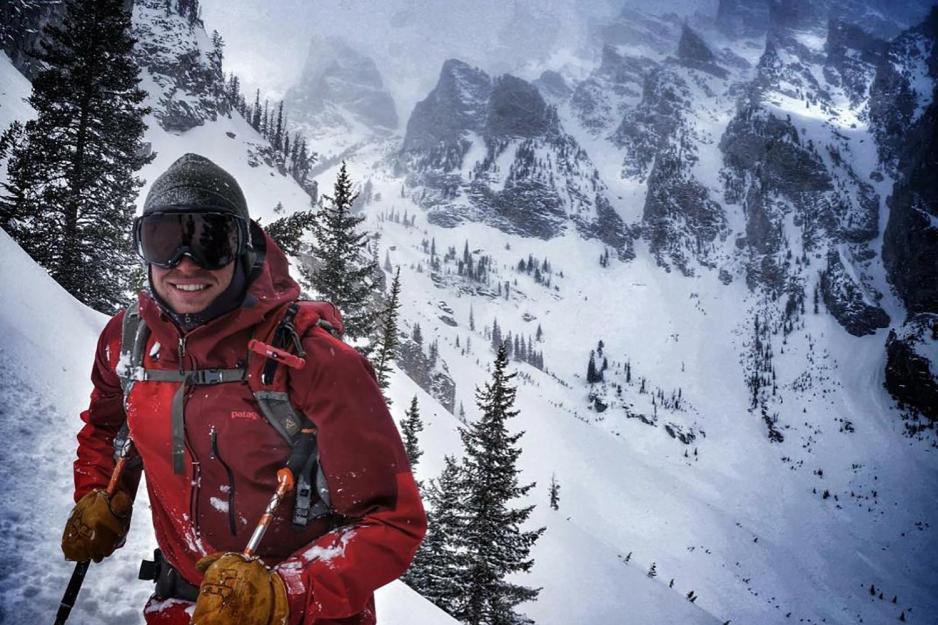 Zach Kuster in a red jacket, ski mask and hat while on a mountain in the snow.  He is looking at the camera