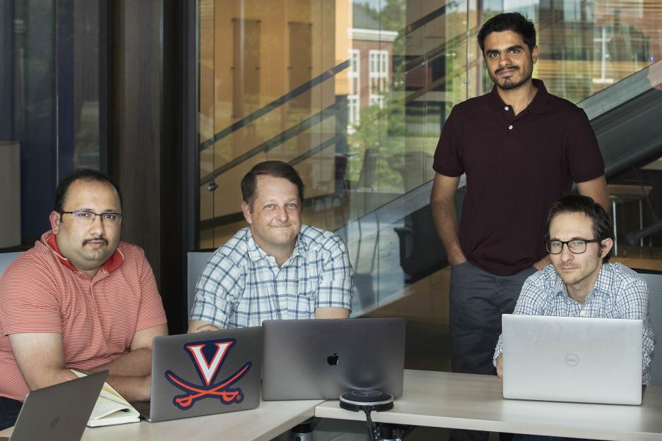 Group of men, 3 sitting with their laptops, and 1 standing behind them all look to the camera