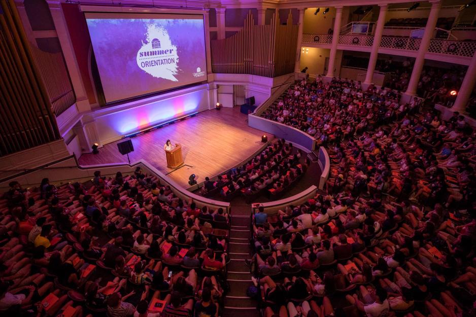A speaker stands at a podium on stage of a Theatre filled with students for Summer Orientation.  