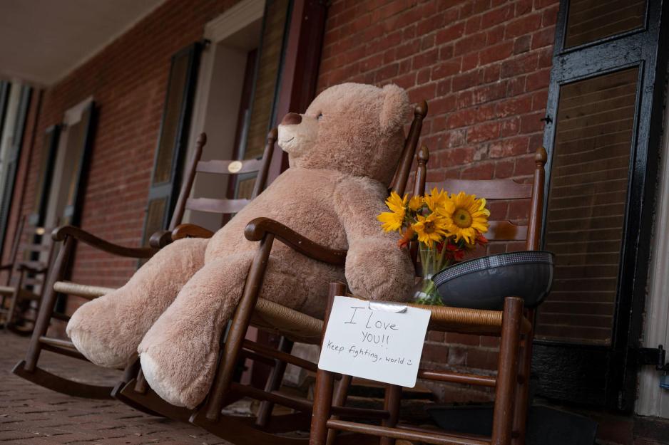 A big stuff brown bear sitting in a wooden rocking chair on the Lawn with flowers next to it and a sign that reads I love you!! keep fighting world