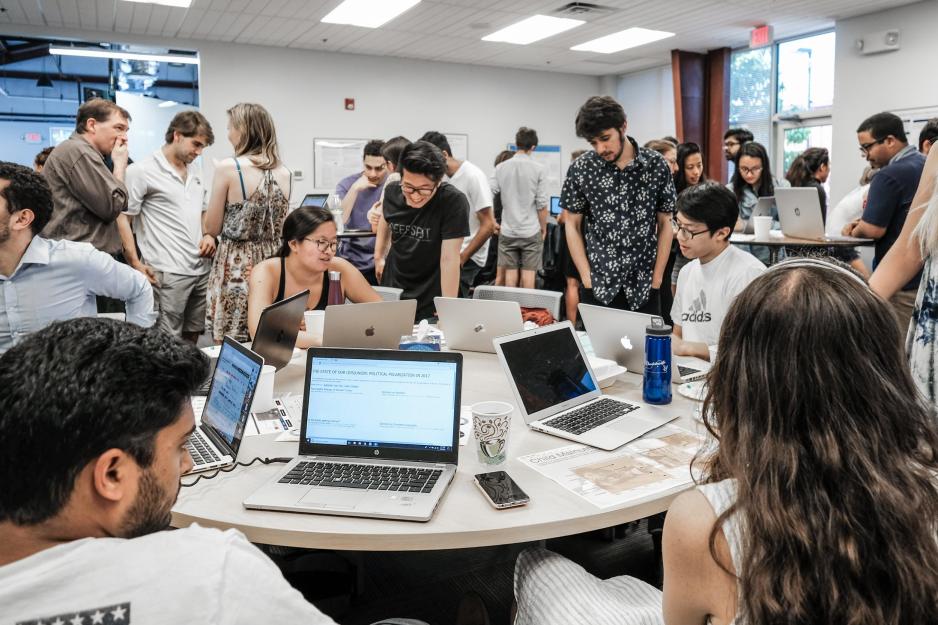 Students stand and sit in a crowded room working on their laptops