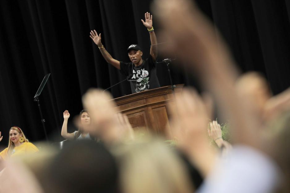 Pharrell Williams speaking at a podium raising his hands as the crowd follows