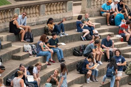 Dozens of students eat lunch and chat in small groups while seated on concrete risers