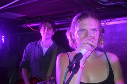 In a darkened basement, a woman sings into a microphone. A man playing bass looks over her shoulder from behind.