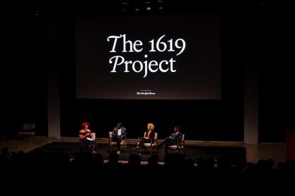 four speakers sit in chairs on a stage speaking with a sign above them that reads The 1619 Project