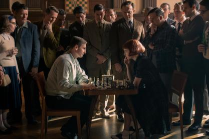 Man and Woman sitting at a chess table staring at each other with a group of people surrounding them.