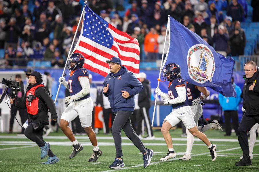 University of Virginia football players run onto the field before a game, one carrying an American flag and another carrying the Virginia state flag. A coach jogs alongside them, and a cameraman films the entrance. Fans in the stands watch in the background on a cool game day.