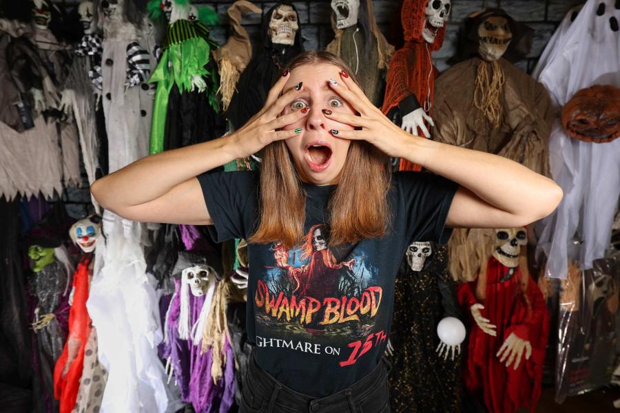 Natasha Bailey candidly posing in a frightened pose in front of a wall of skeleton scary figurines. On her shirt is says ”Swamp Blood, Nightmare on 15th” and depicts a woman with skull makeup casting a spell with a snake around her neck.