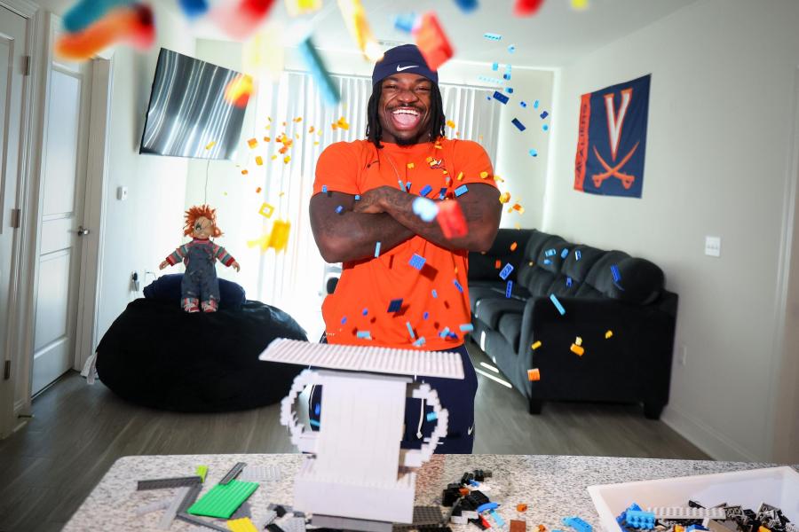 J’Mari Taylor laughing with his arms crossed in an apartment front room surrounded by confetti. The room has a V-Saber flag on the wall, a Chucky doll in the background and several legos are on the table in front of Taylor.