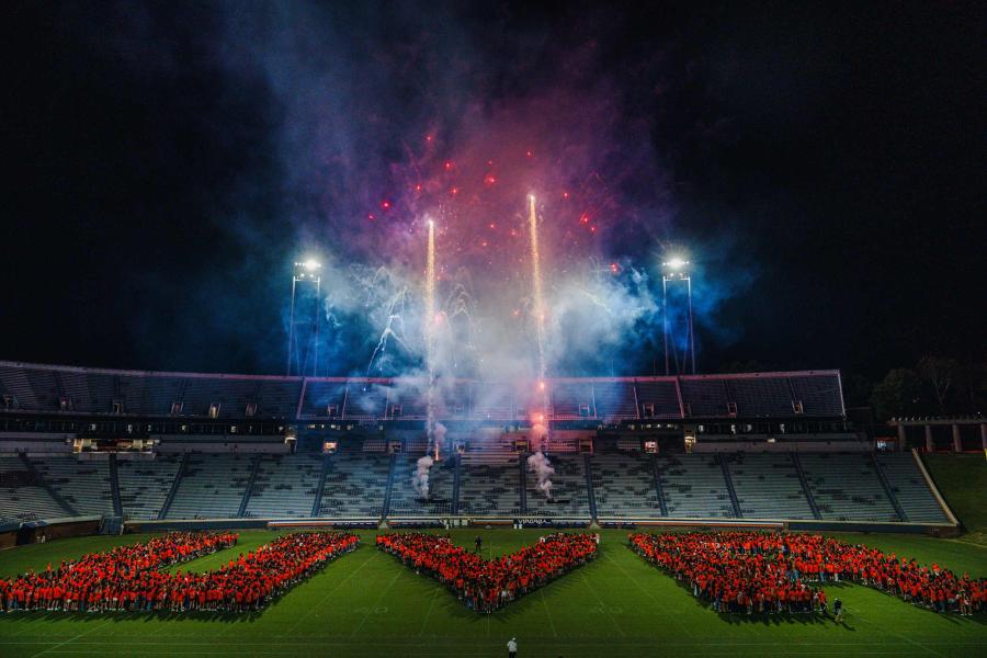 several people spell out the letters UVA on the field of Scott Stadium with fireworks in the background