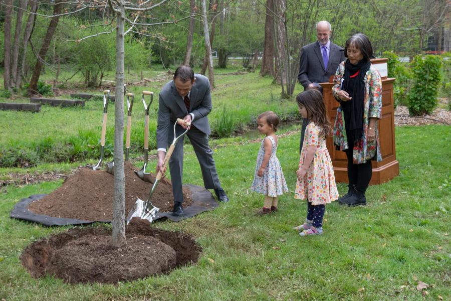 Someone shovels dirt at the base of a freshly planted tree as a family watches