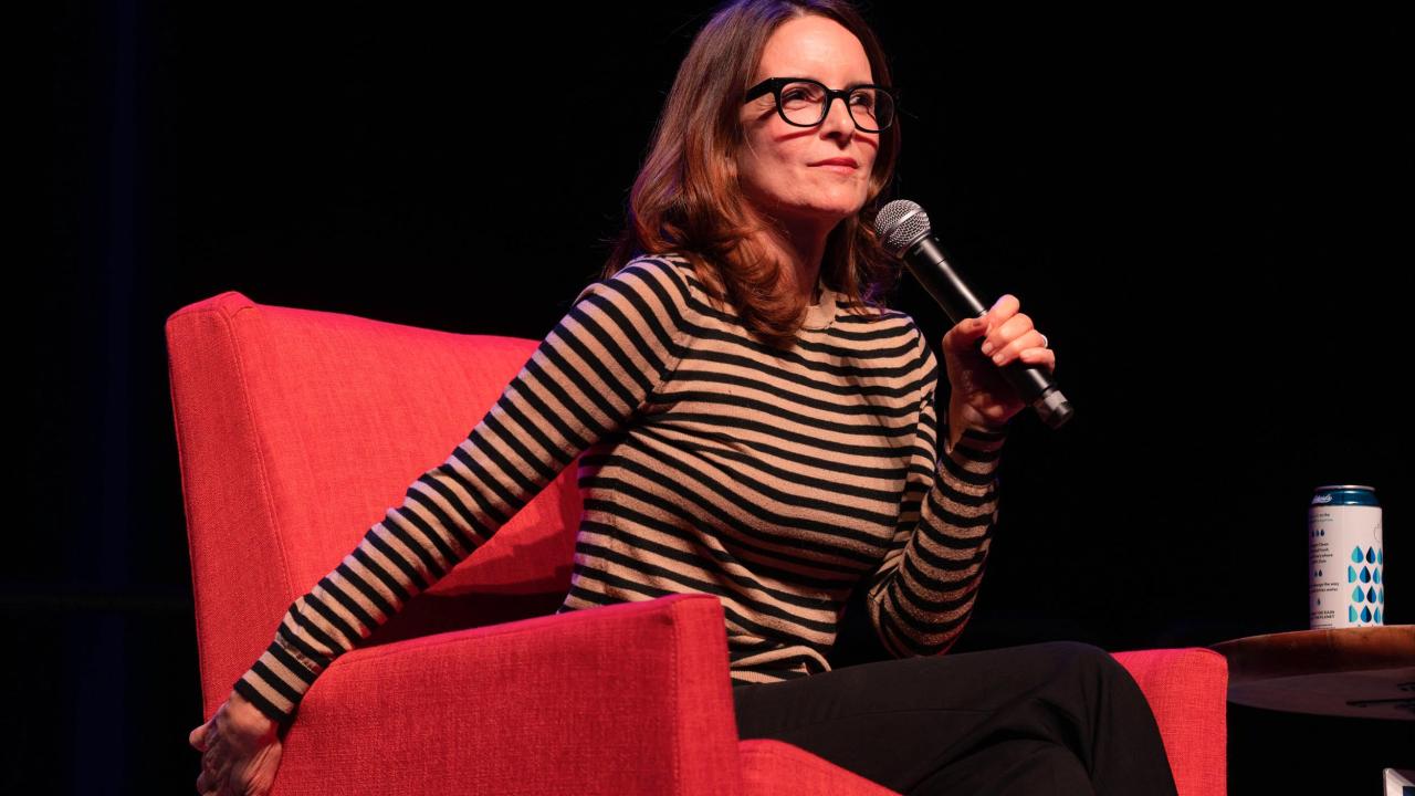 Candid photo of Tina Fey on stage sitting in a red chair holding a microphone