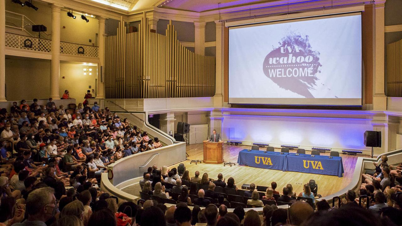 Students sitting in chairs on the lawn looking at a stage where a woman is giving a speech