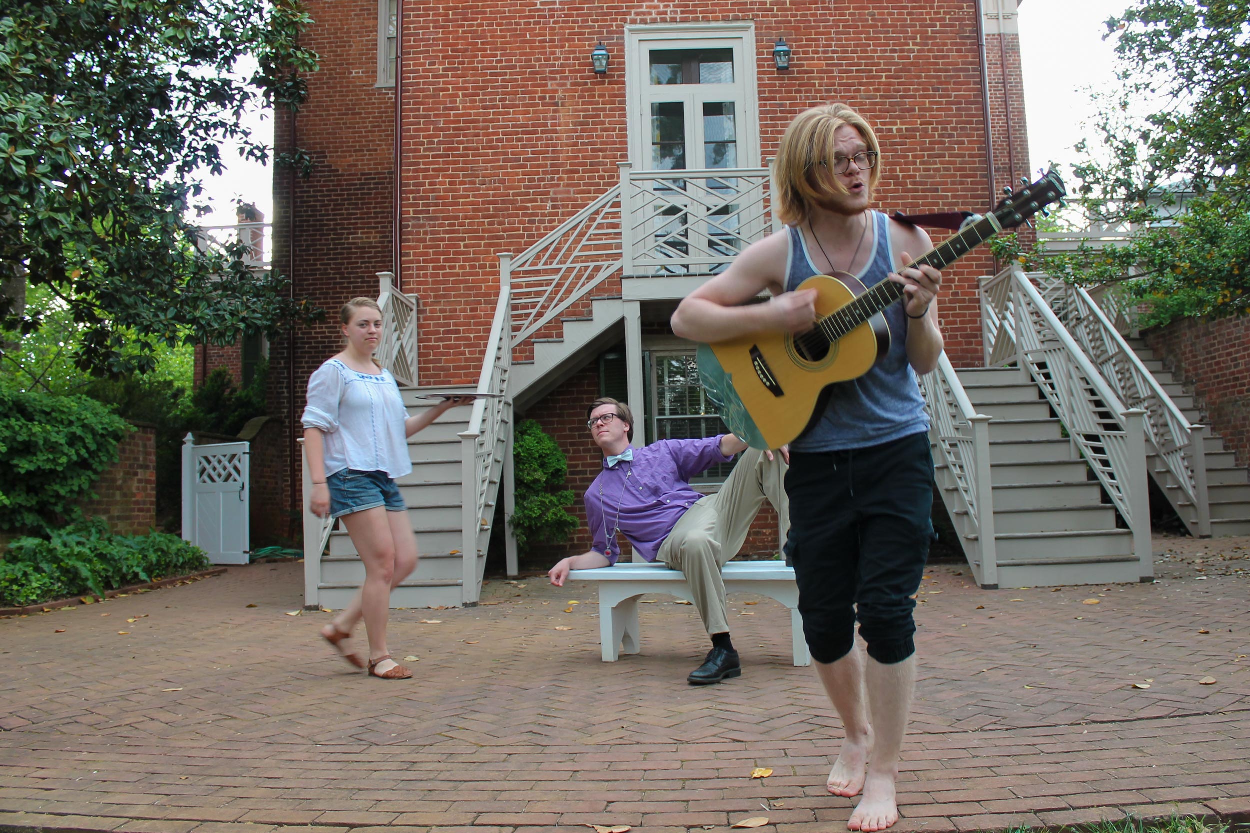  Annika Schunn, Pete Hanner and Nick Hurst outside of a building playing a guitar and acting