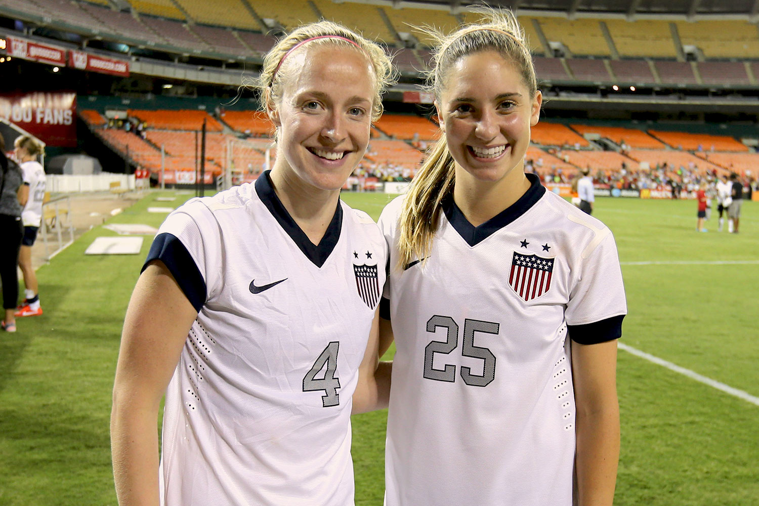 Two USA female soccer players stand together with arms around each other smiling to the camera