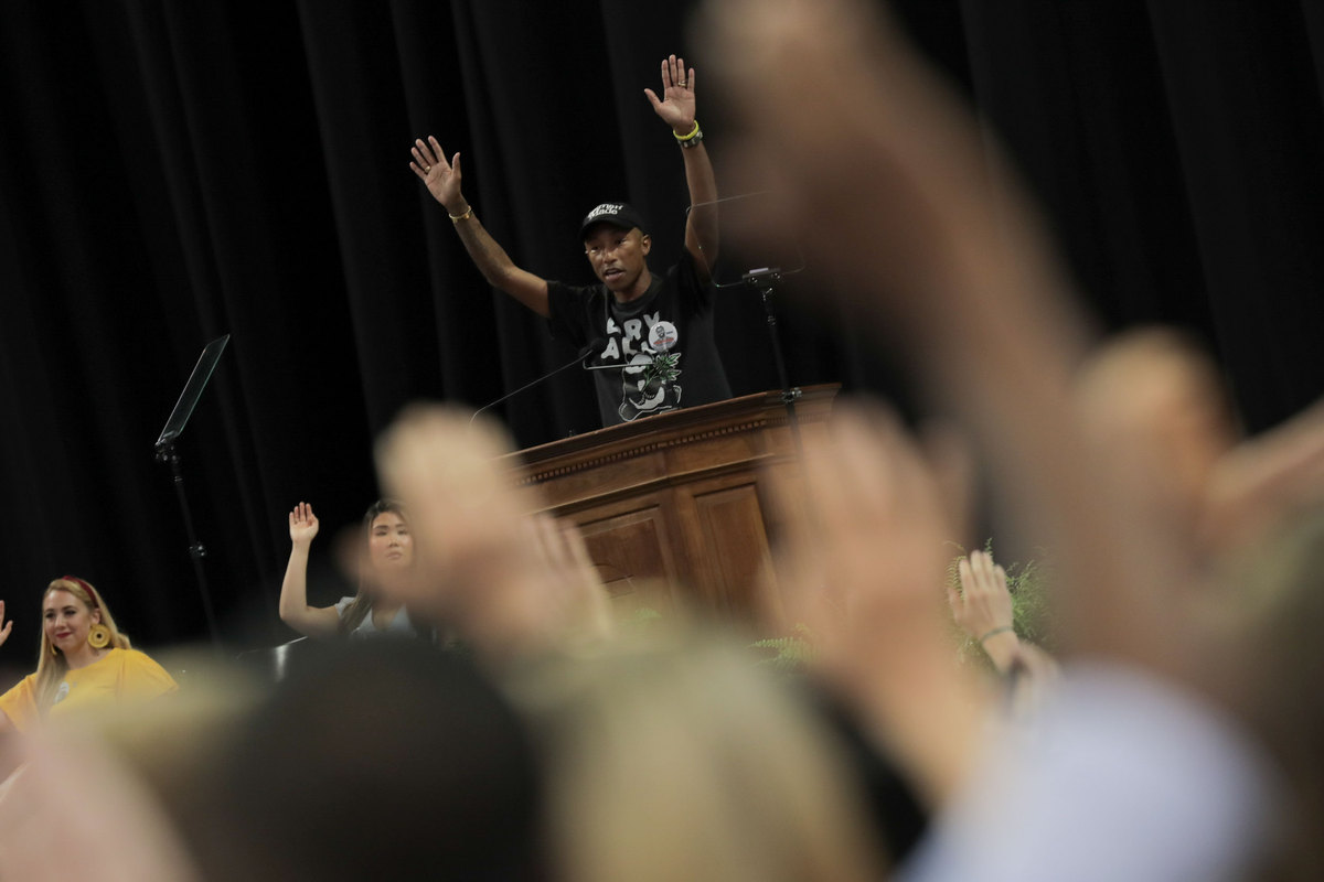 Pharrell Williams speaking at a podium raising his hands as the crowd follows