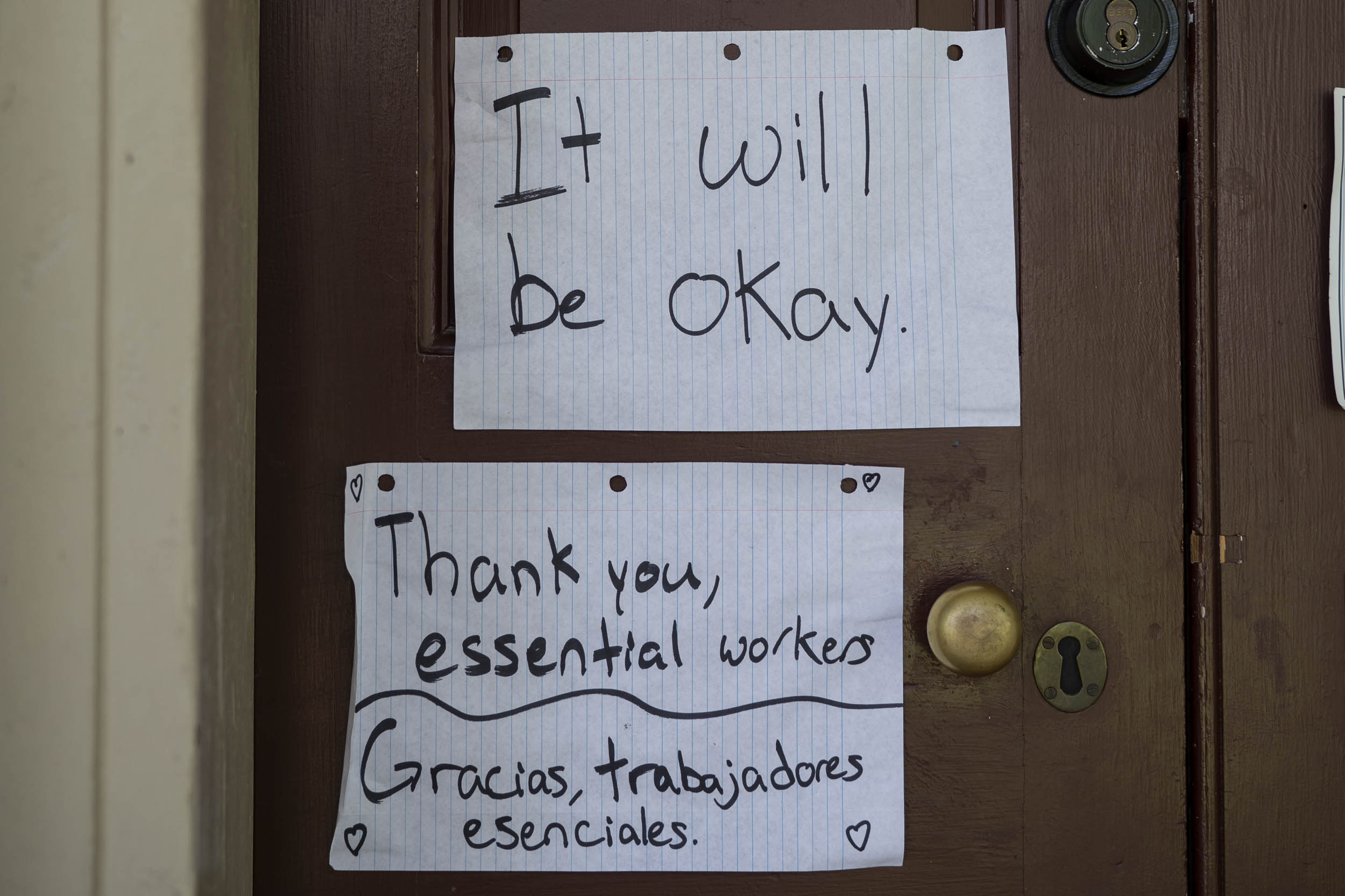 signs on looseleaf paper attached to a brown door that read: It will be okay. thank you, essential workers, Gracias, trabajadores esenciales