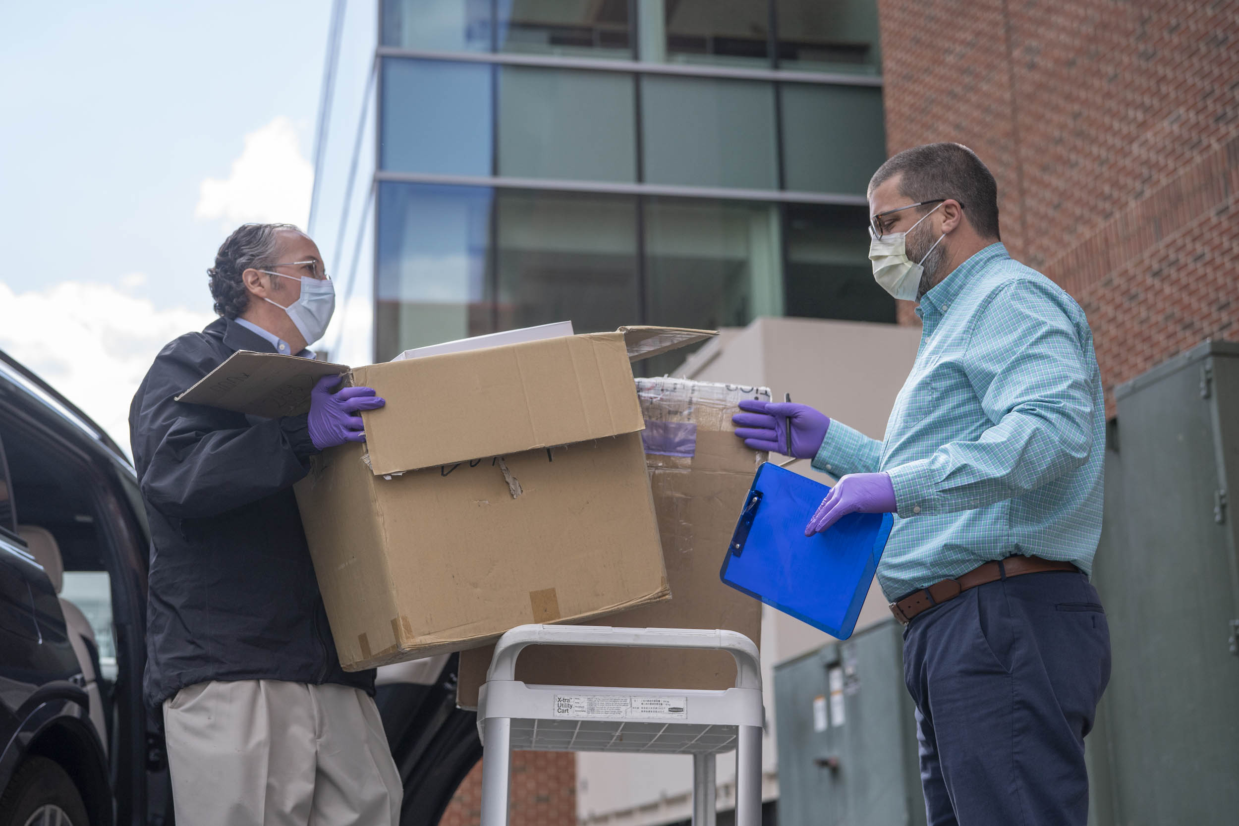 Two men placing cardboard boxes on a trolly.  One man (left) is getting the boxes out of the car the man (right) is watching as he holds a clipboard