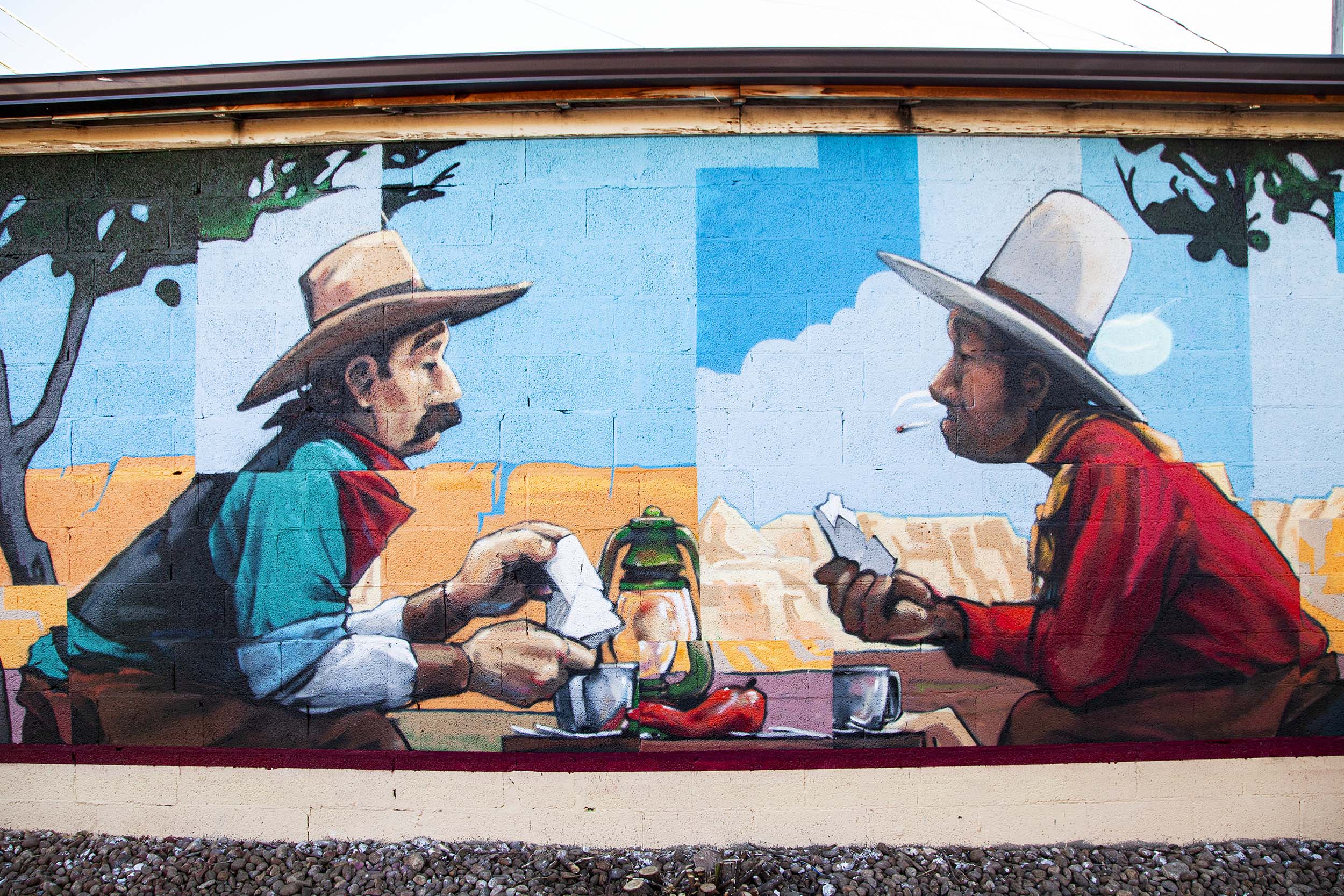 painting on a wall of two hispanic men sitting at a table together with food starting at each other in the face