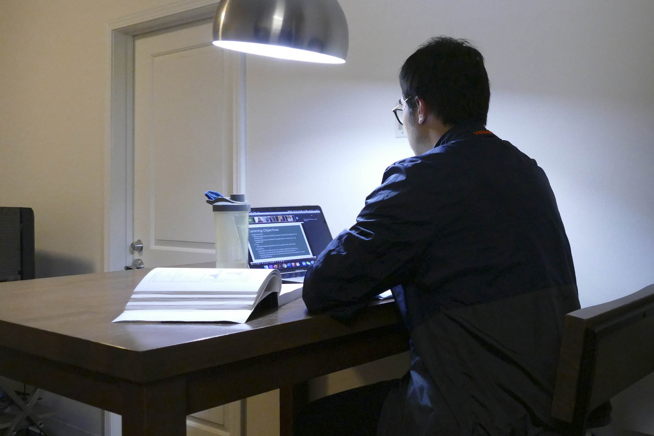 Winston Tang sits at a table with a desk light over head working on his computer with an open book on the desk