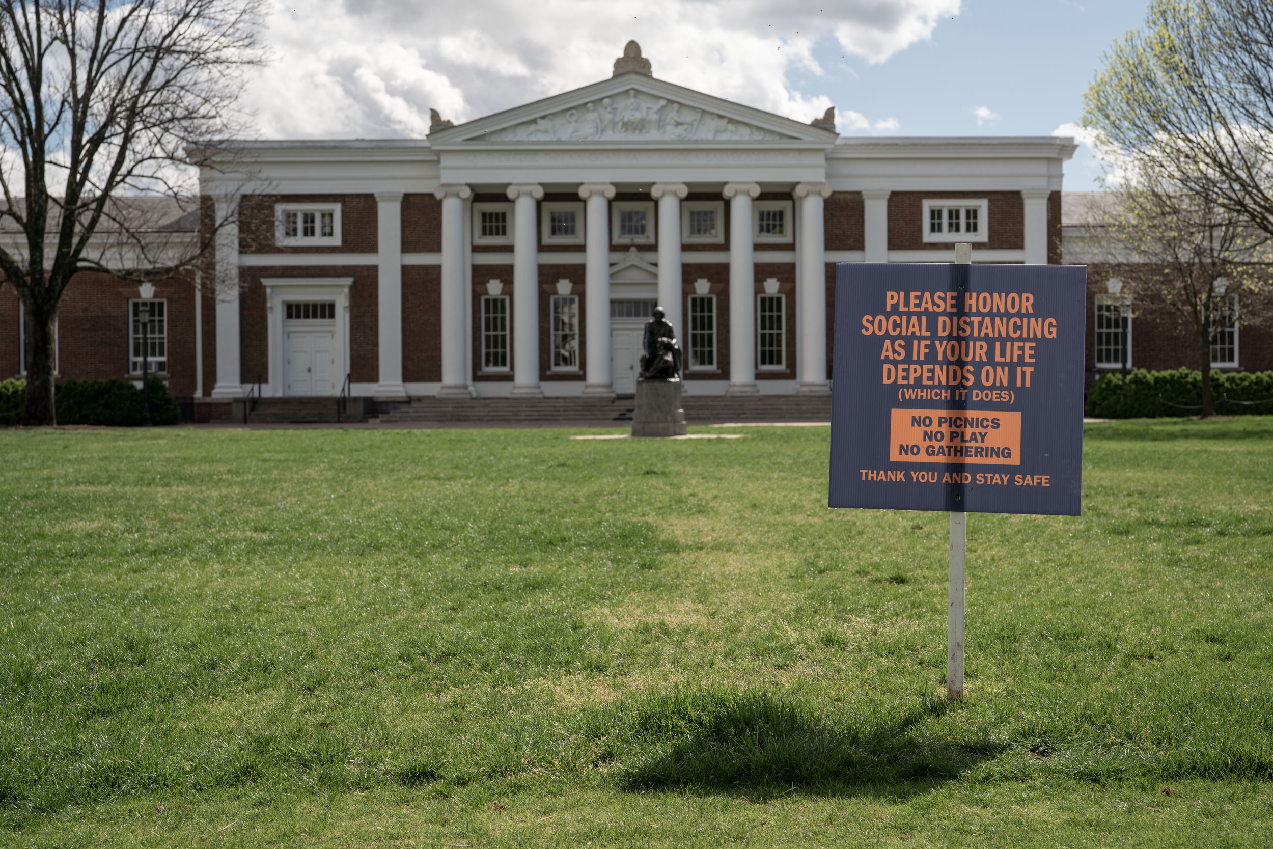UVA building with a sign in the grass that reads, please honor social distancing as if your life depends on it (which it does) no picnics, no play, no gathering, thank you and stay safe.